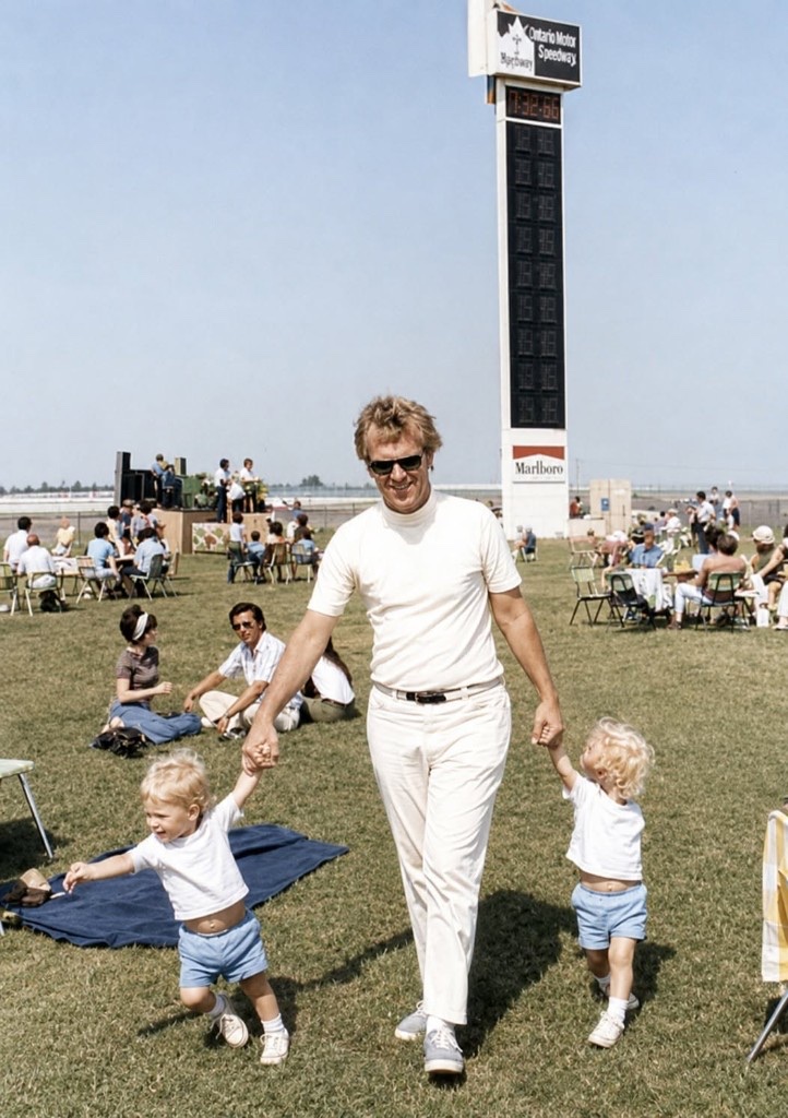 Monty Roberts holding hands with twin toddler boys on the grass at Ontario Motor Speedway, with the Ontario Motor Speedway scoring pylon, timer 7:32.66, and Marlboro branding behind them